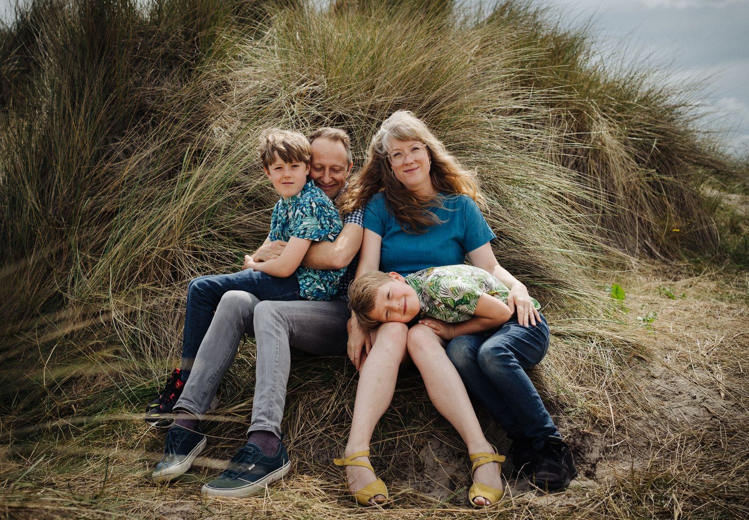 Family of four sitting together among tall grass dunes, smiling outdoors on a cloudy day.