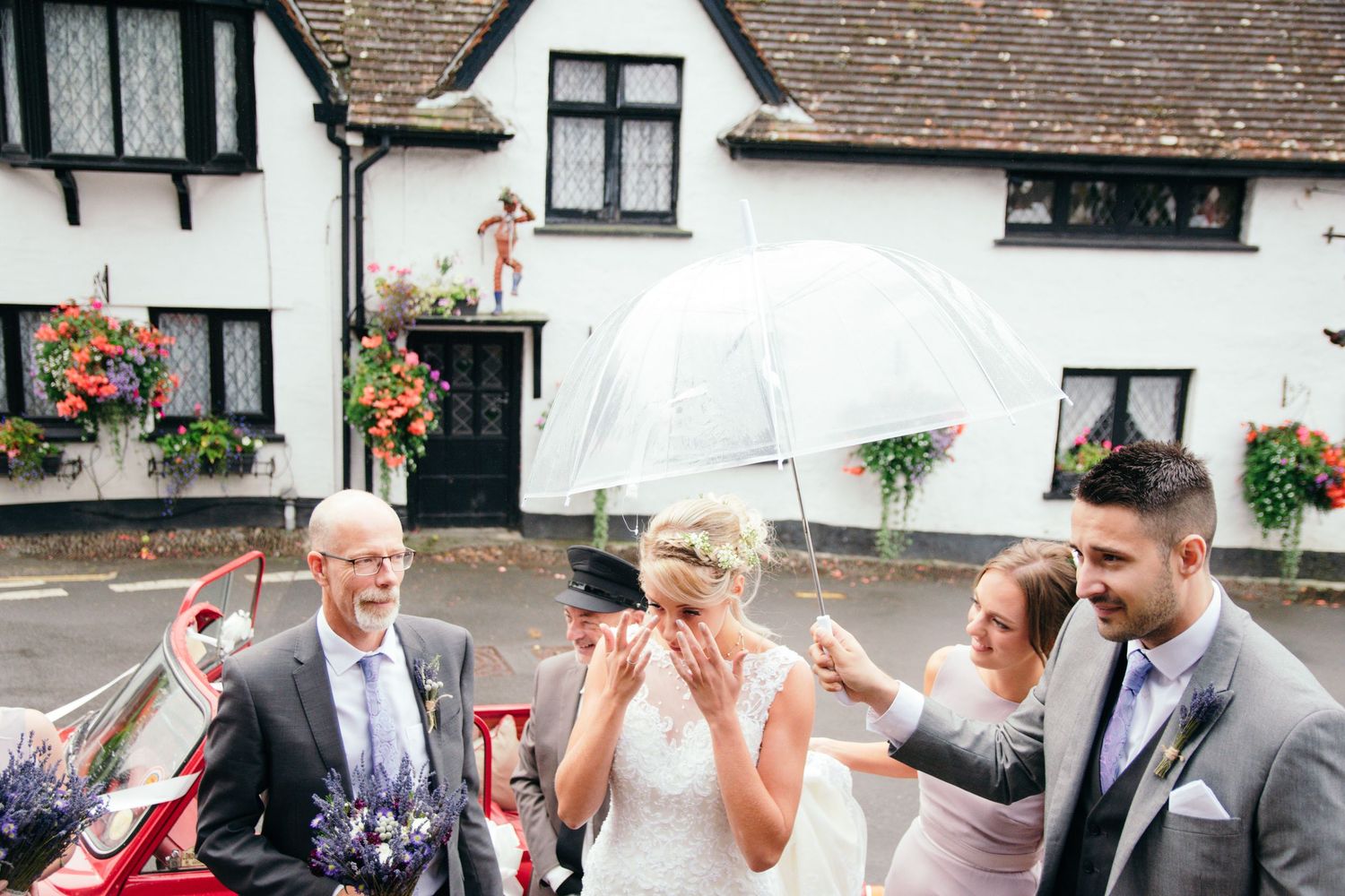 Bride laughing under clear umbrella with wedding party outside white English pub on a cloudy day.