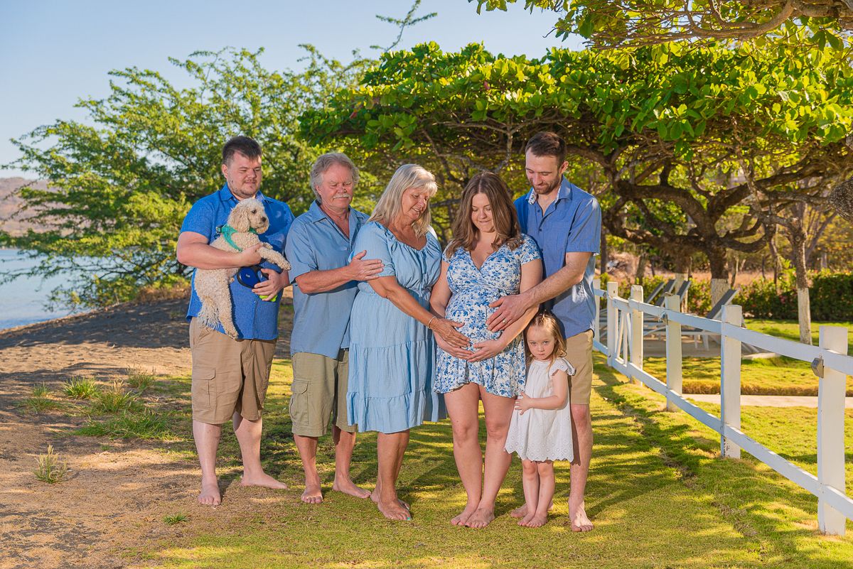 Multigenerational family maternity photo outdoors on green grass with white fence, trees, and ocean view in Hawaii.