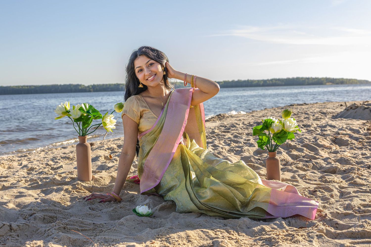 Woman in green saree sitting on sandy beach with flower vases, smiling by the water at golden hour.