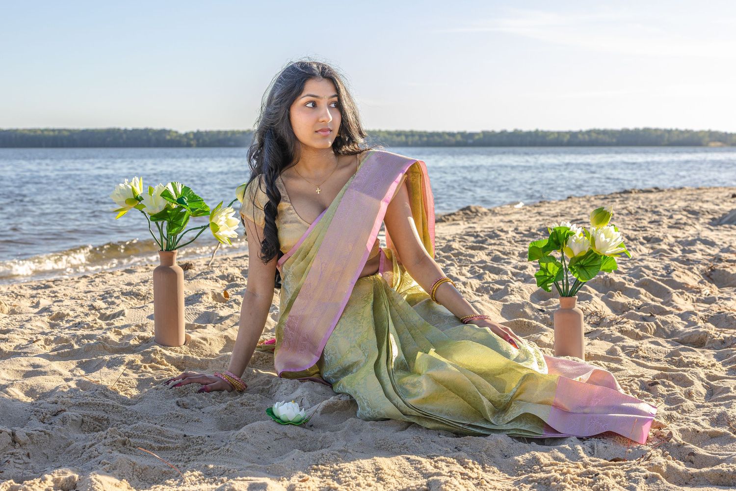 Young woman in green and pink saree sitting on sandy beach with flower vases at sunset.