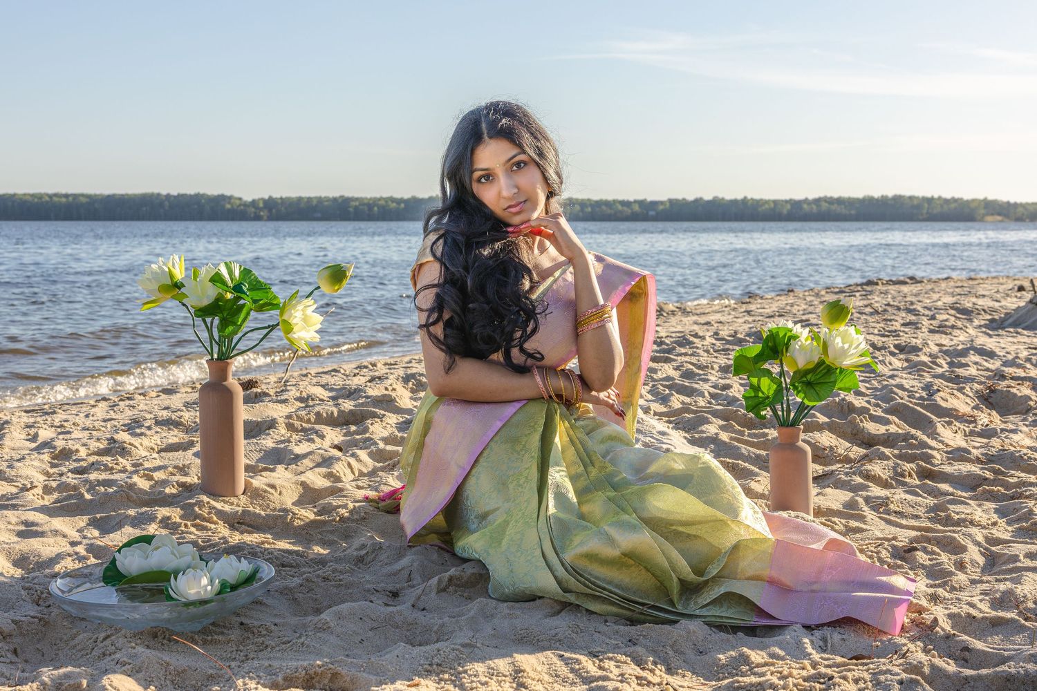 Young woman in green saree sitting on sandy beach with lotus flowers in vases during golden hour.