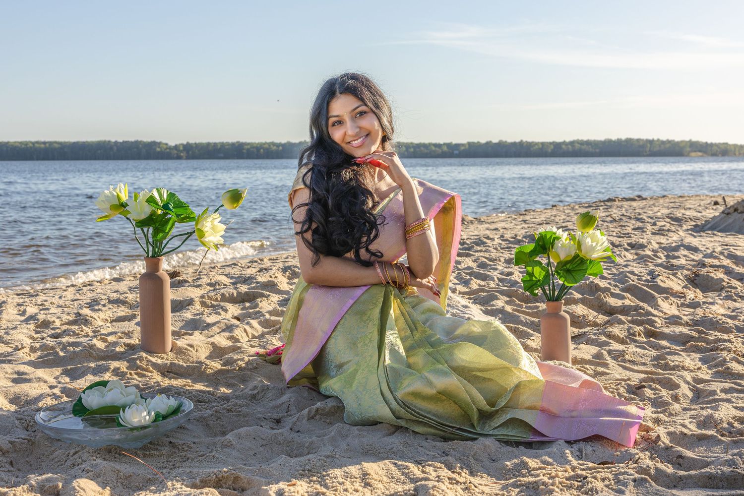 Smiling woman in elegant green saree posing on beach surrounded by white lotus flowers at sunset.