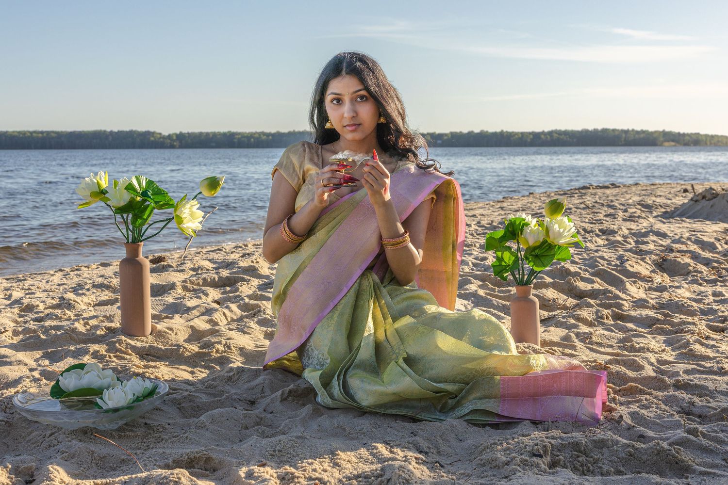 Woman in green and pink saree sitting on beach sand with lotus flowers in vases at sunset.