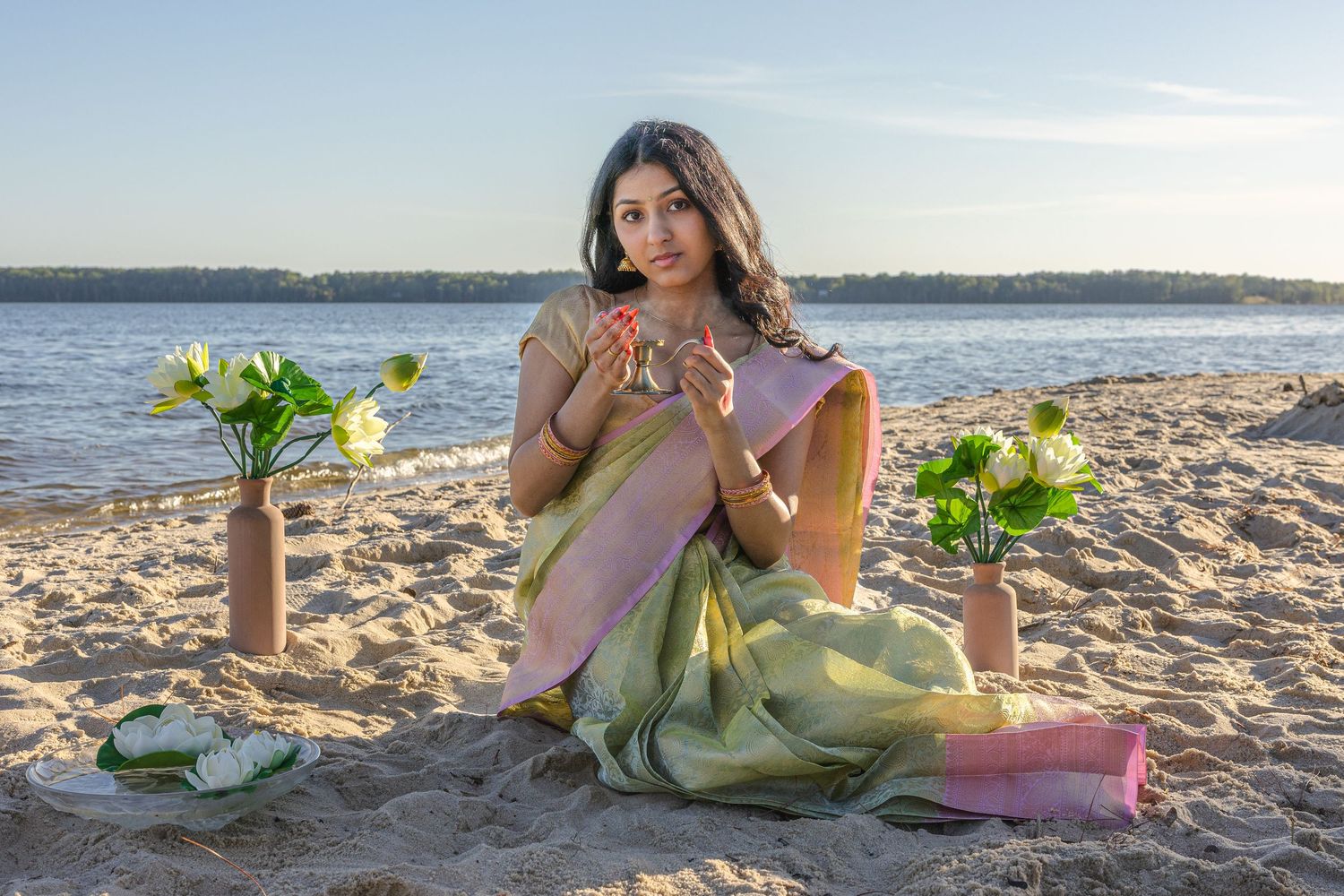 Woman in traditional green and pink saree holding lotus flowers while sitting on sandy beach.