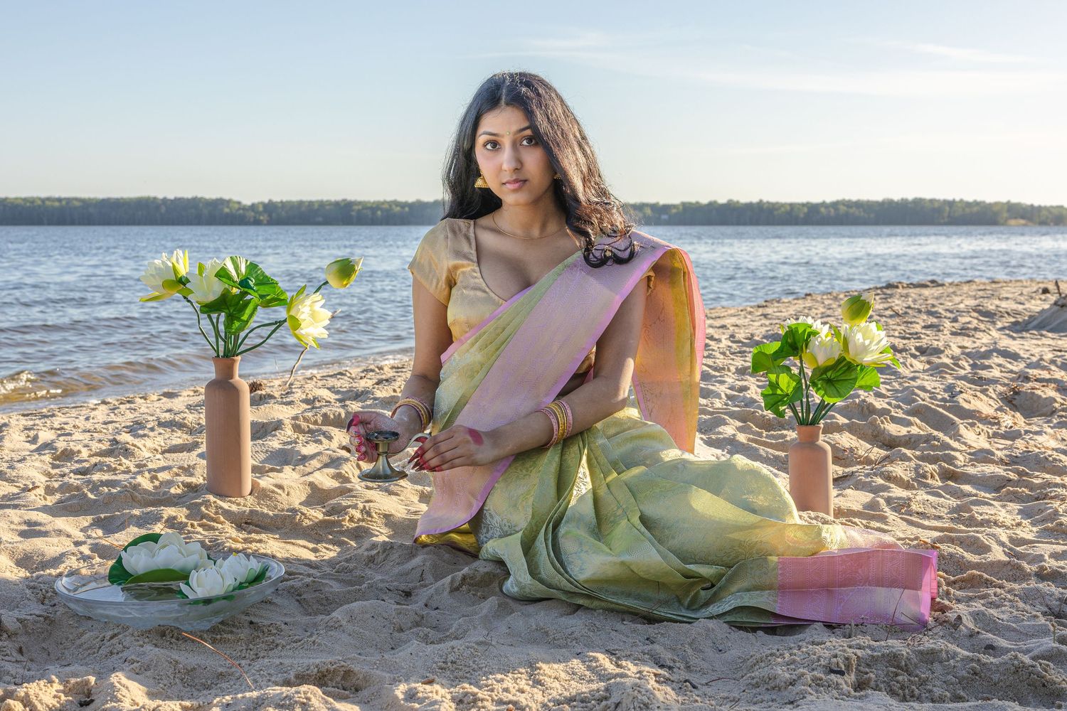 Woman in green and pink saree sitting on beach sand with flower vases during golden hour.