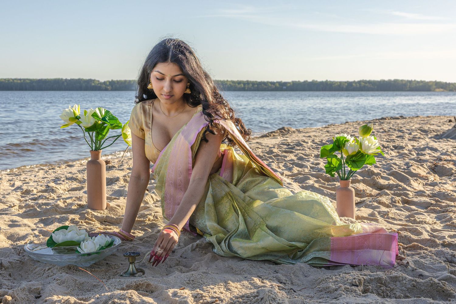 Woman in green saree sitting on sandy beach with lotus flowers in vases during golden hour.