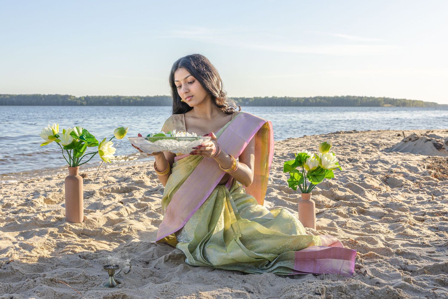 Young woman in traditional green saree holding floral arrangement on sandy beach at sunset.
