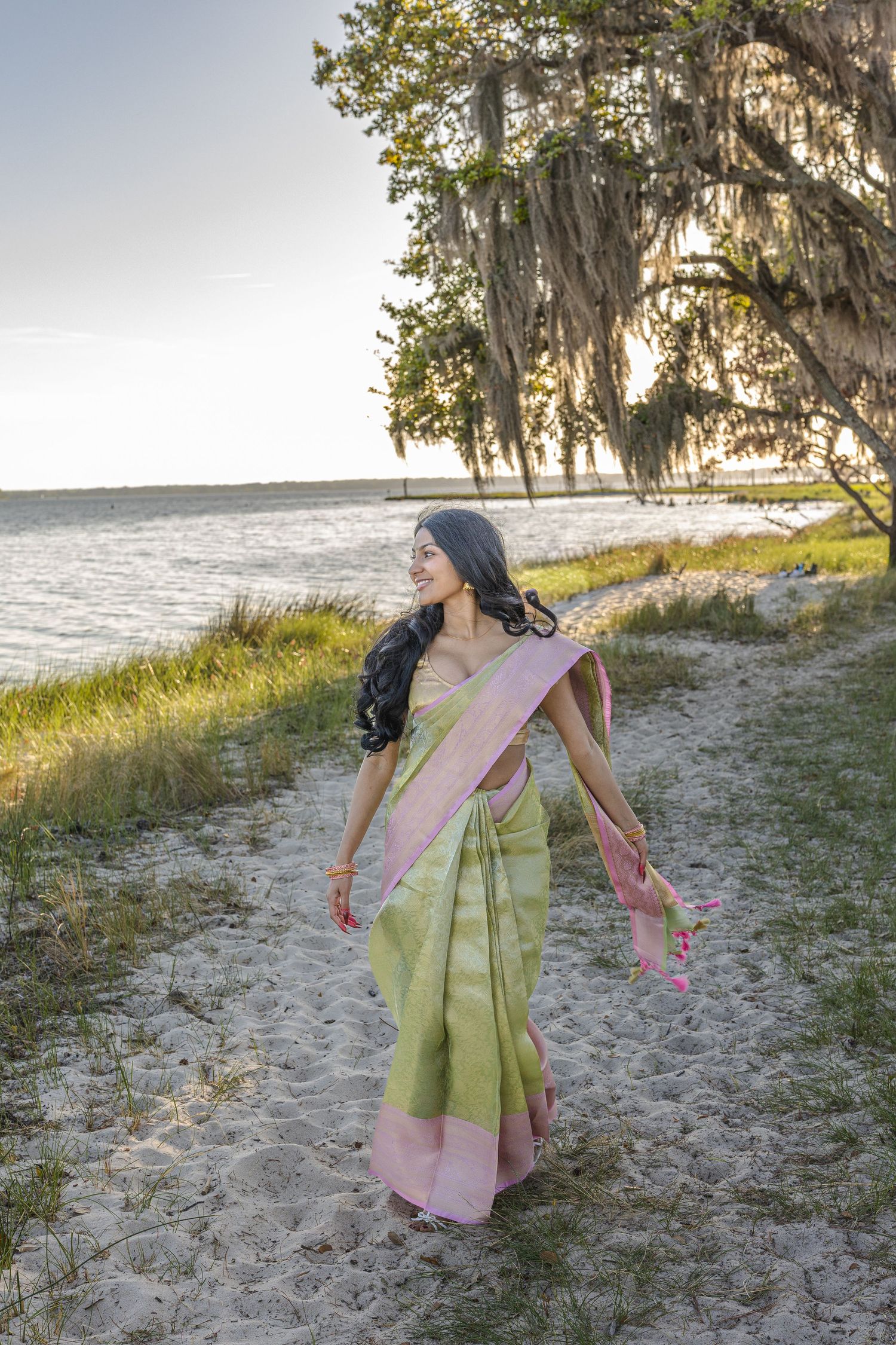 Woman in traditional green pink saree walking on sandy beach path near water under mossy oak tree.
