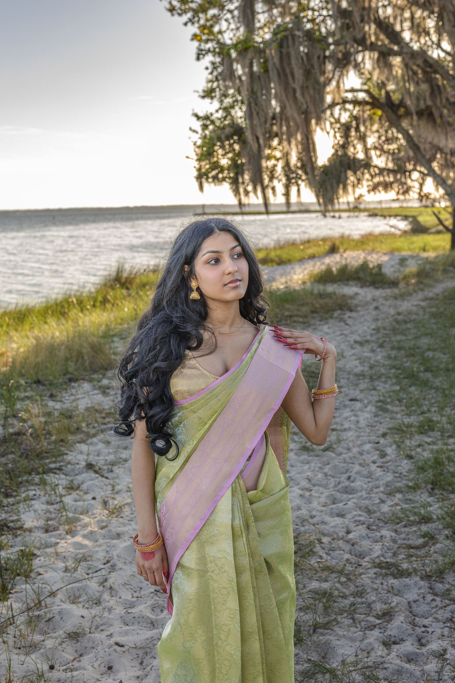 Young woman in green and pink saree poses on sandy shore near water and moss-draped oak tree at sunset.