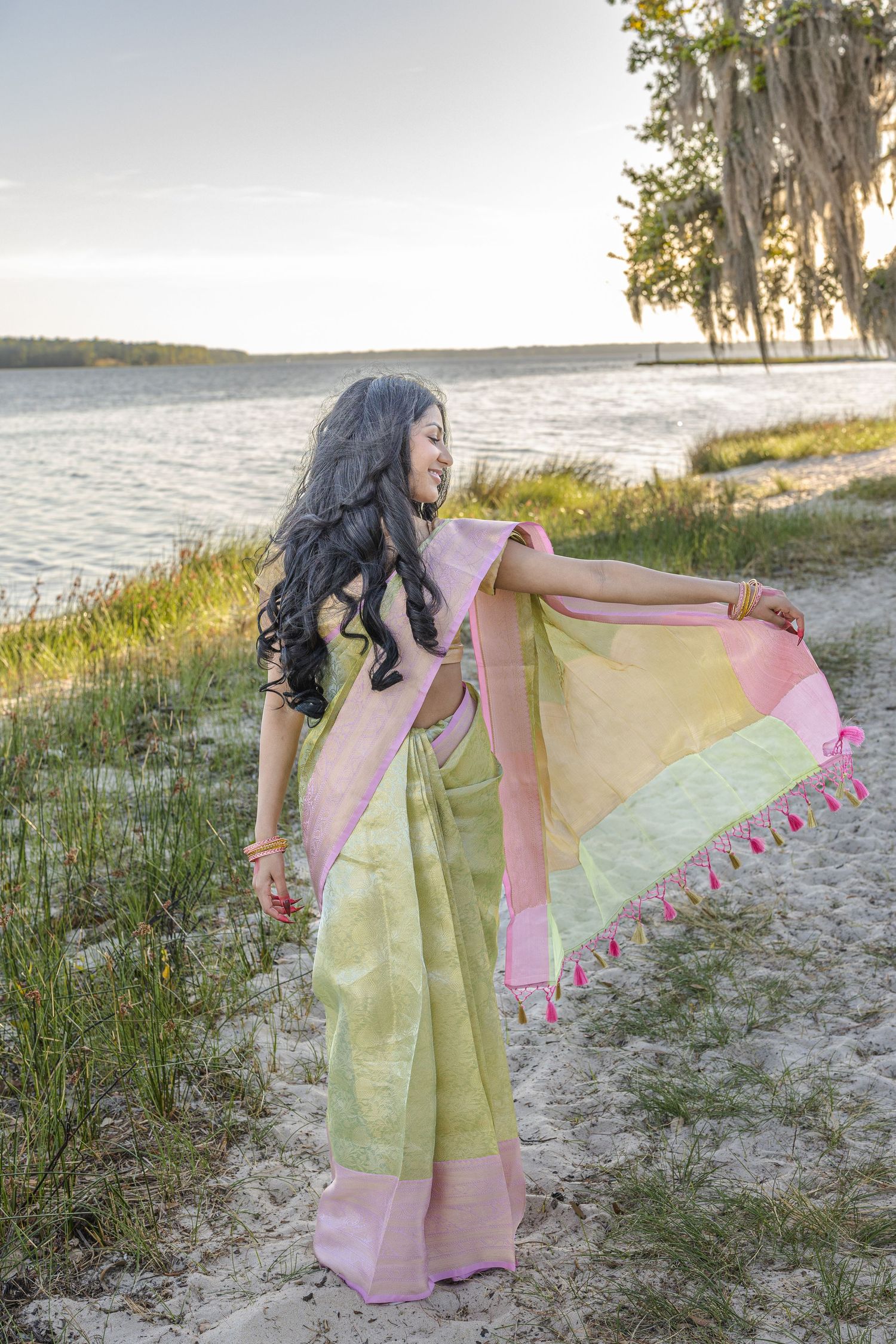 Woman in green pink saree with tassels posing on sandy beach near water at golden hour sunset.