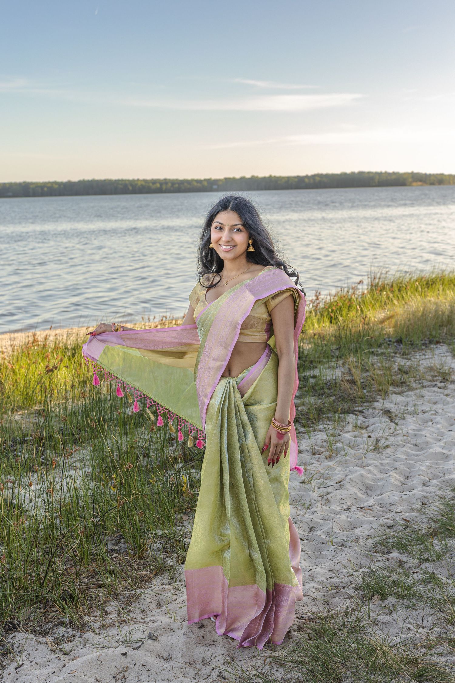 Young woman in green and pink saree poses gracefully on sandy beach at sunset with water in background.