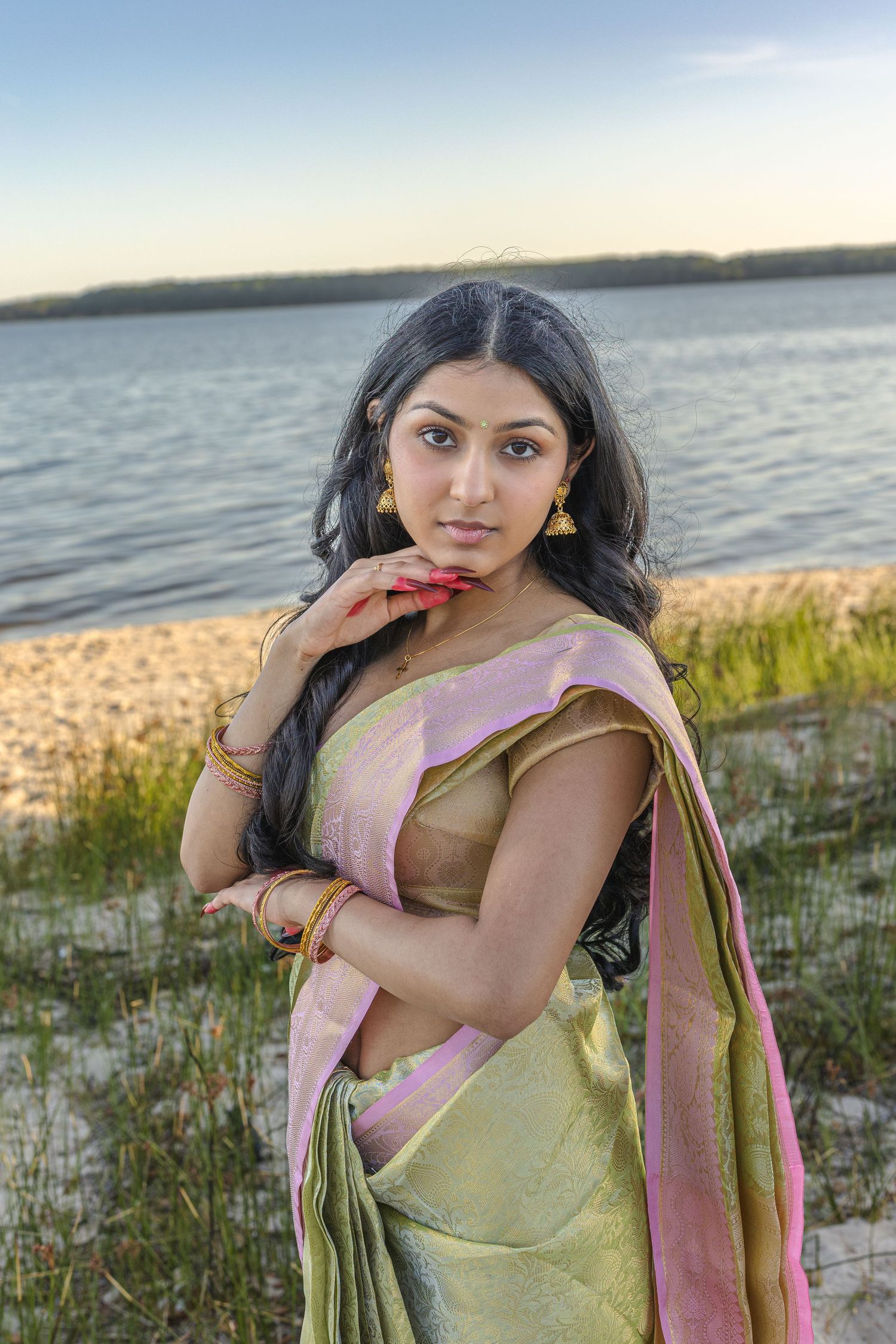 Beautiful woman in traditional green and pink saree posing on beach at golden hour.