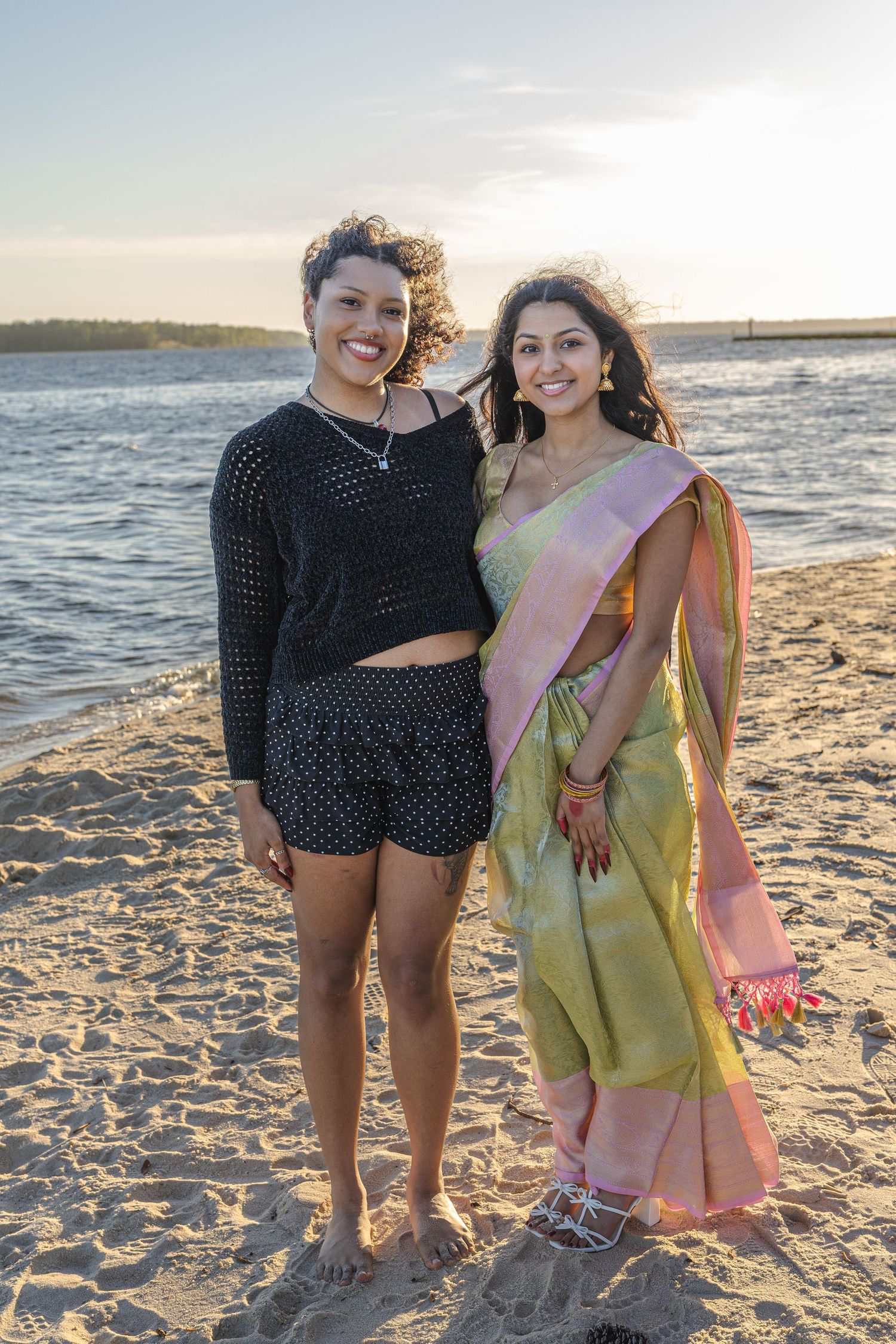Two women smiling on a sandy beach at sunset, one in a black outfit and one in a traditional green and pink saree.
