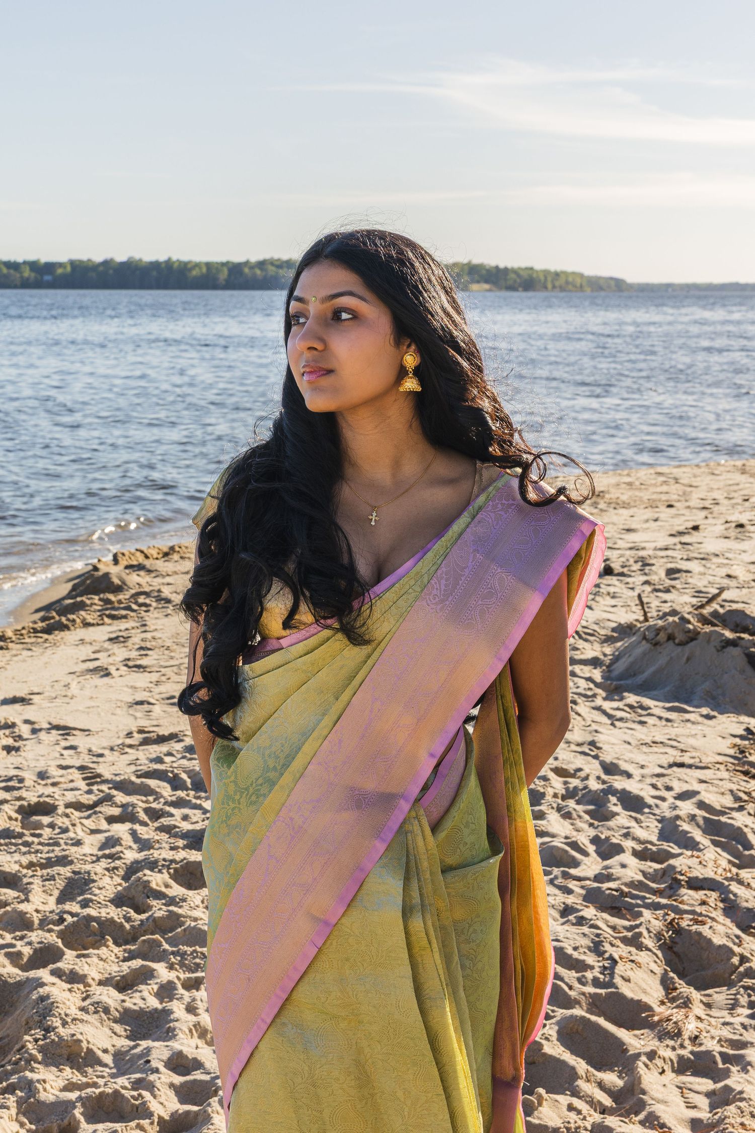 Beautiful woman wearing traditional green and pink saree standing on sandy beach with river backdrop.