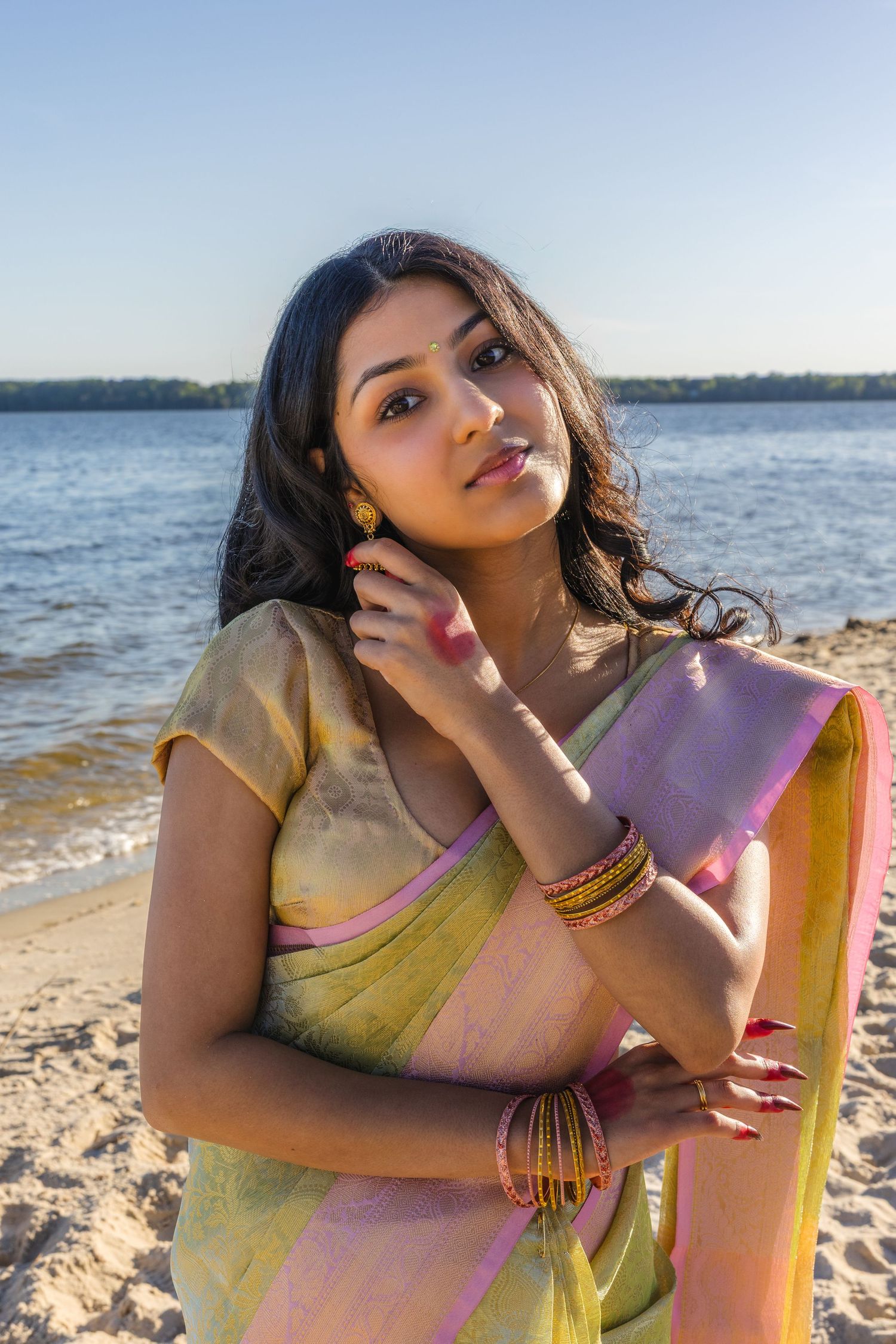 Young Indian woman in traditional green and pink saree with gold bangles posing gracefully on a sandy beach.