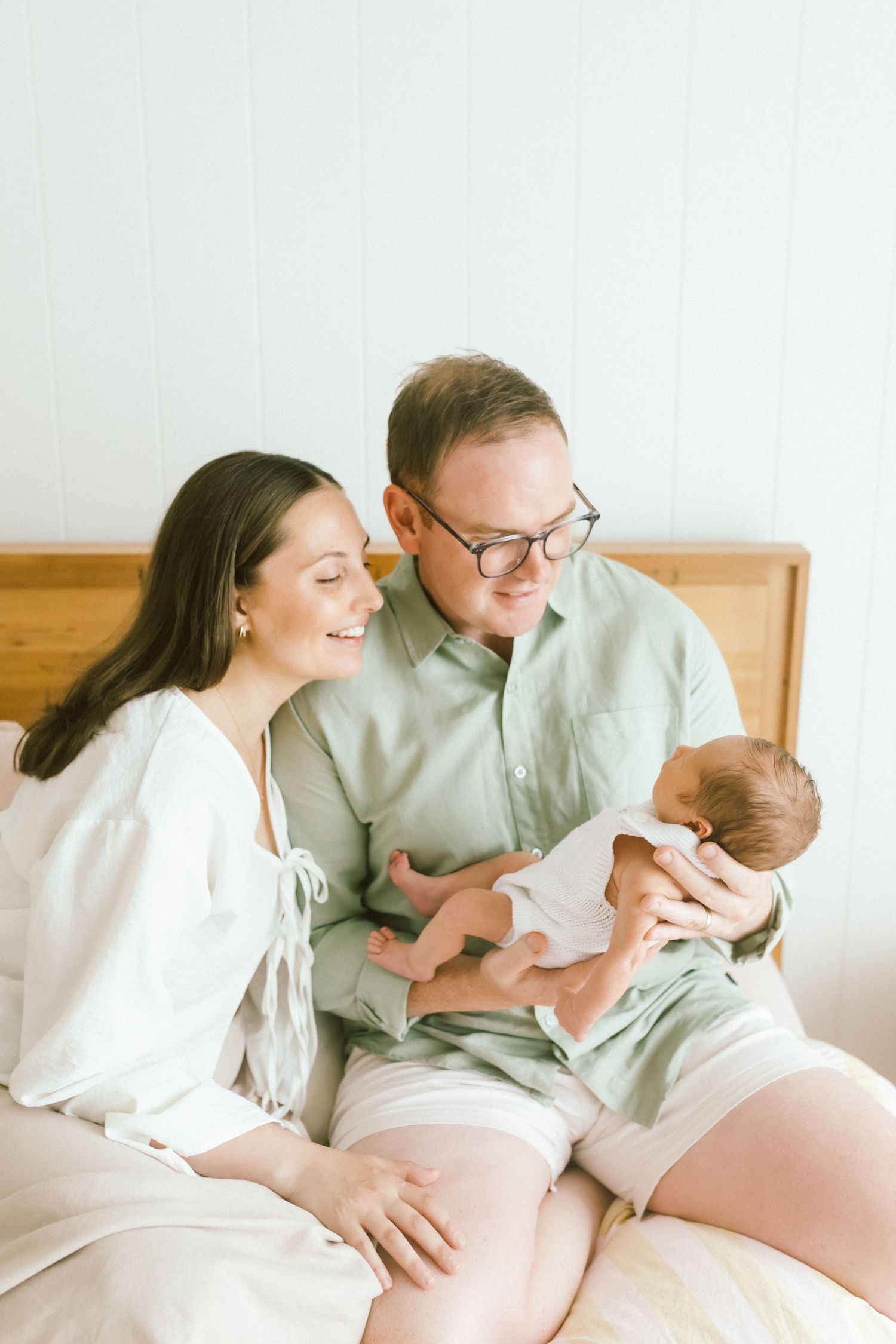 Happy parents sitting on bed, lovingly gazing at their newborn baby in a bright, airy room.