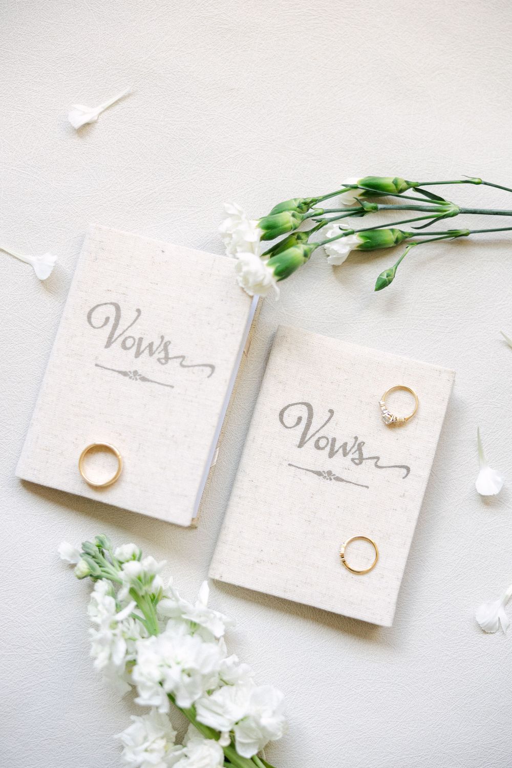 Two elegant vow books with gold wedding rings and white flowers on a soft white background.