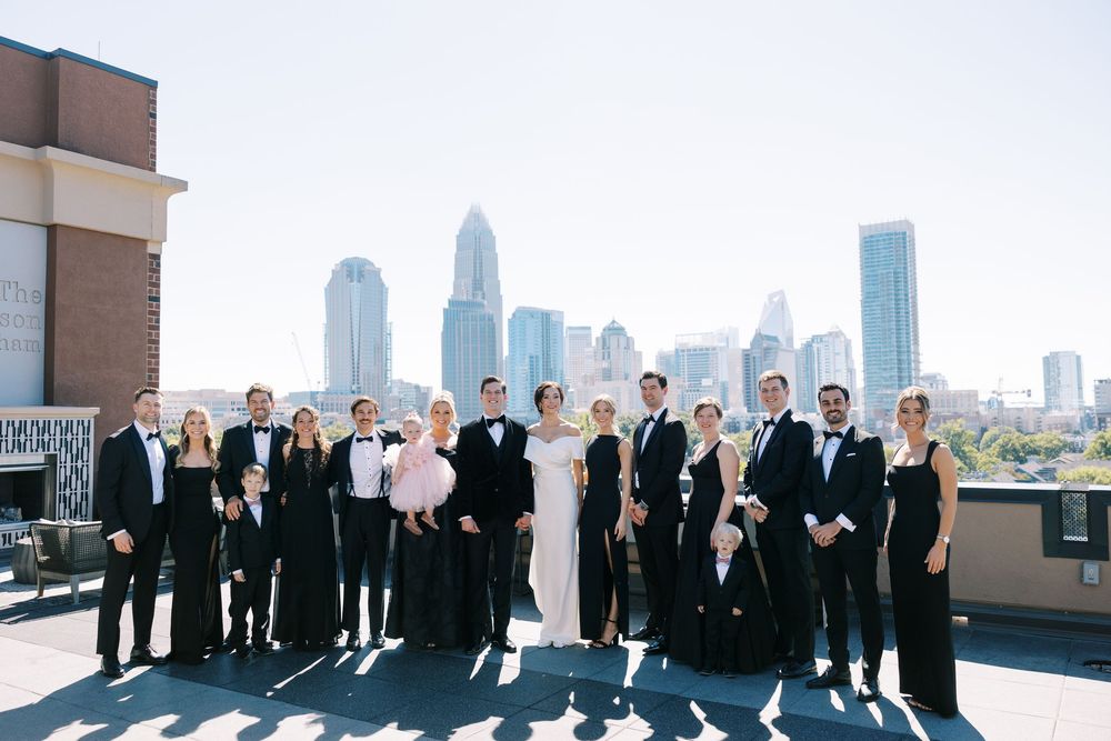 Wedding party in black formal attire poses on rooftop with Charlotte NC city skyline backdrop on sunny day.