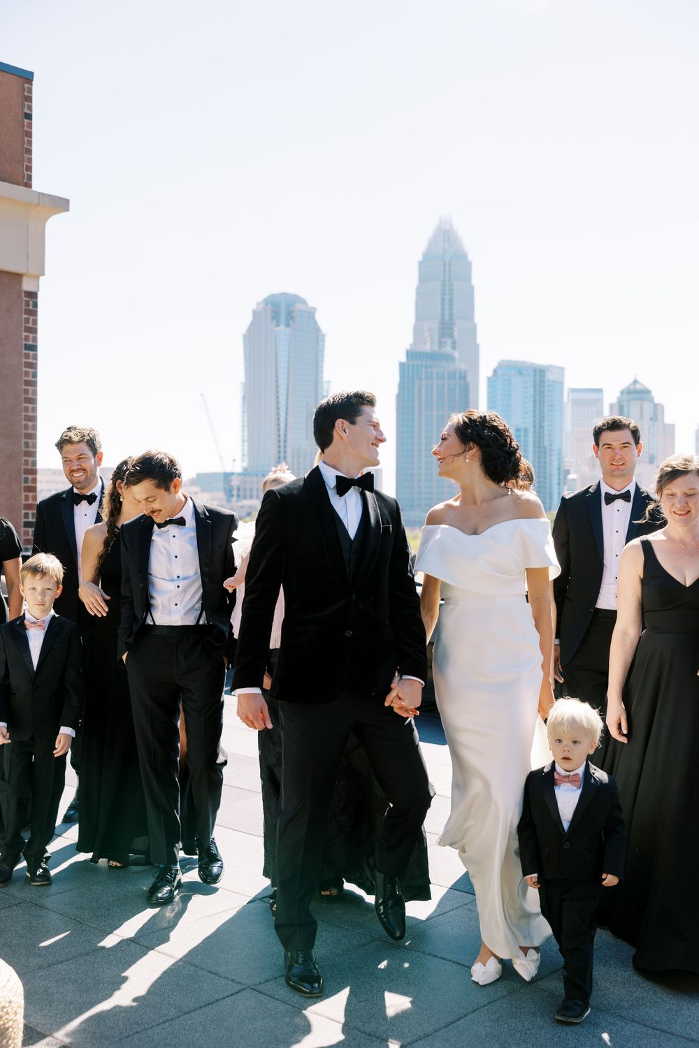 Wedding party walks on rooftop with city skyline backdrop, bride in white off-shoulder gown, groom in black tuxedo.