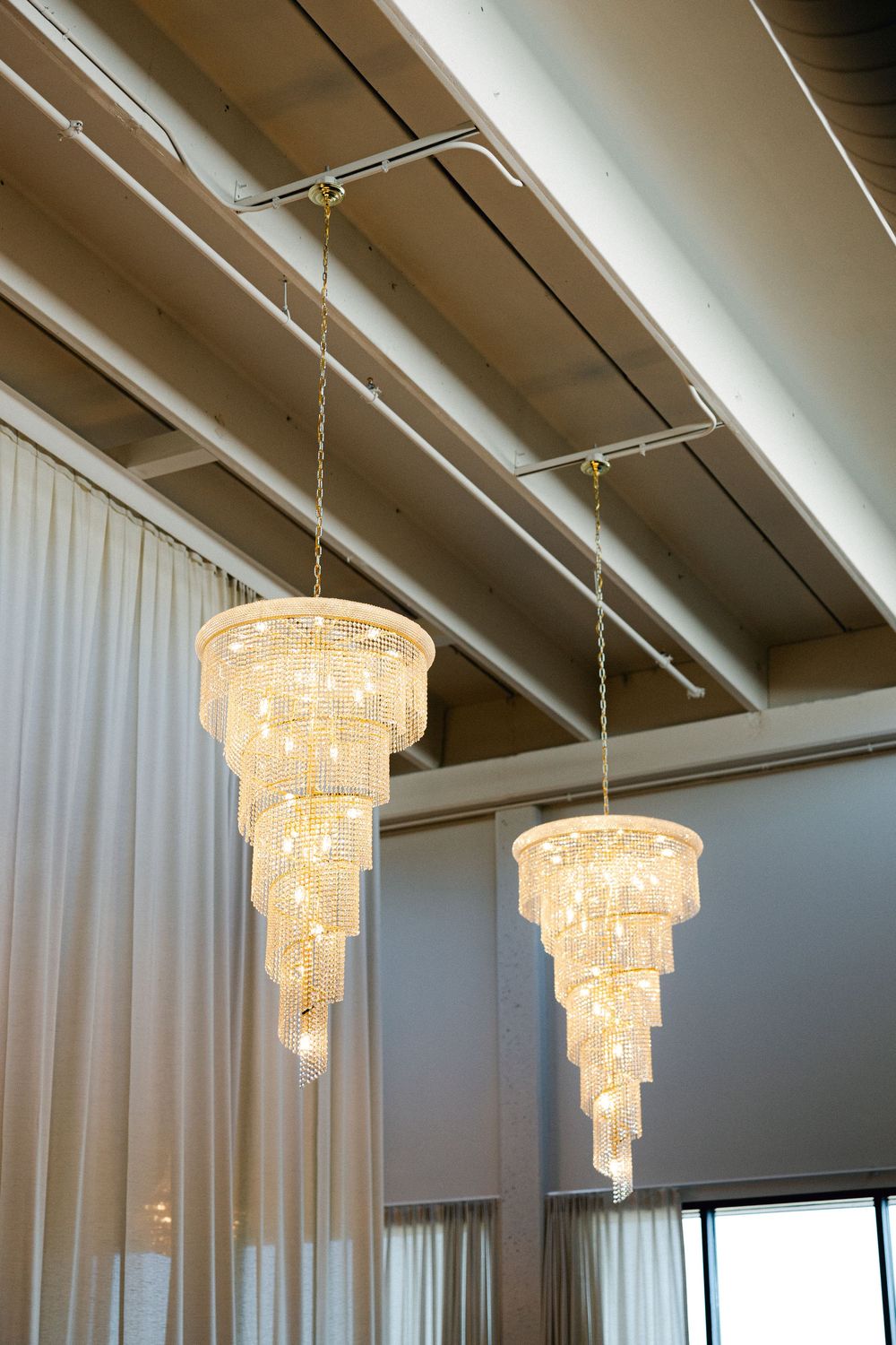 Two elegant tiered crystal chandeliers hanging from a modern ceiling with exposed beams and white curtains.