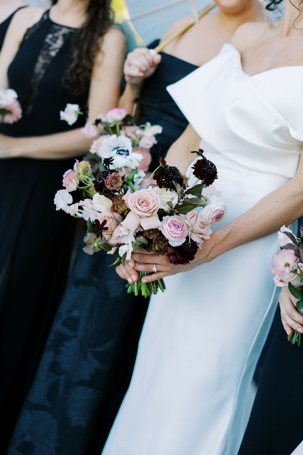 Bride in white gown holding romantic bouquet of blush roses and dark blooms, with bridesmaids in black dresses.
