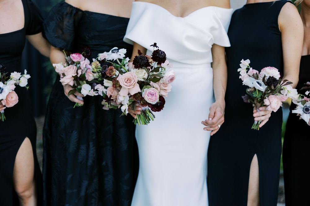 Bride in white gown with bridesmaids in black dresses holding pink and burgundy floral bouquets.