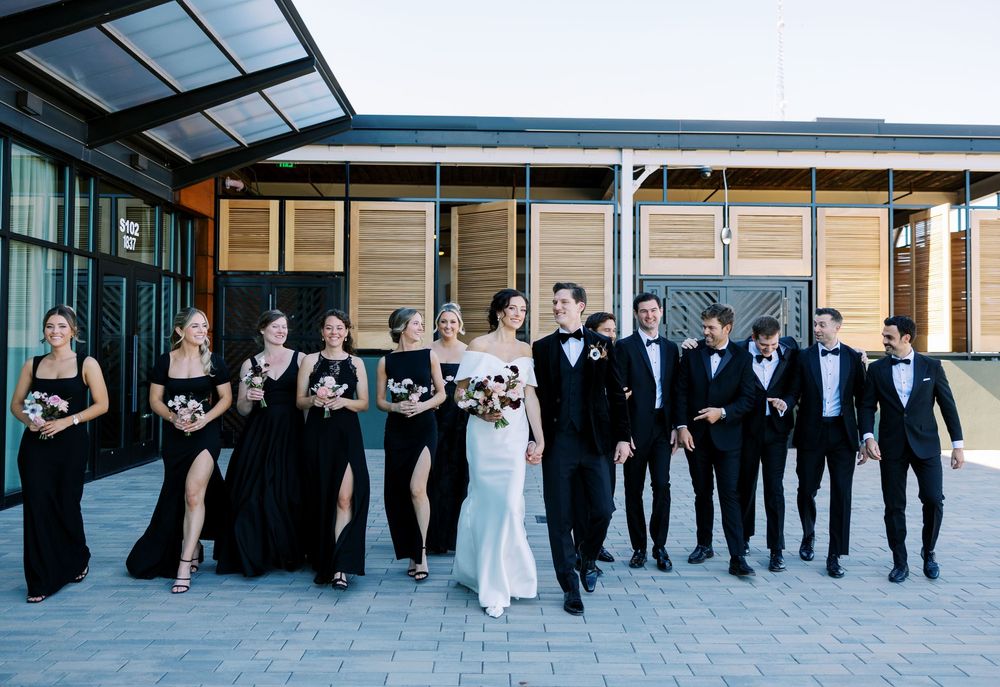 Wedding party in black attire walks together outdoors, bride in white gown holding bouquet, groom in tuxedo.