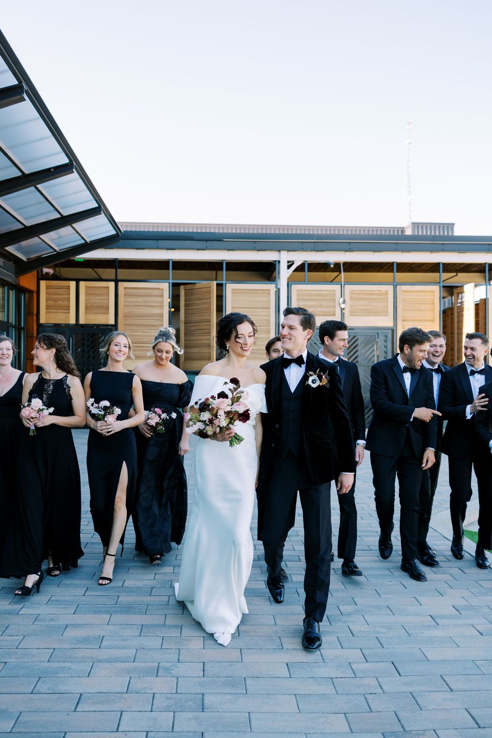 Bride and groom walk with wedding party in black attire on paved outdoor venue path.