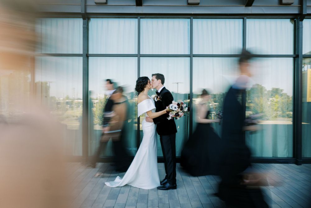 Bride and groom share a kiss while wedding party moves around them in a modern venue.