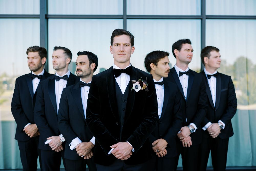 Groom in black velvet tuxedo stands before groomsmen in matching black tuxedos and bow ties at a wedding.