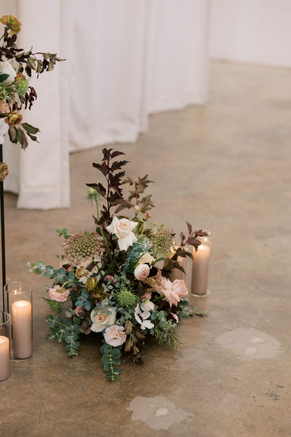 Elegant wedding floor arrangement with blush roses, green blooms, and candles on concrete floor.