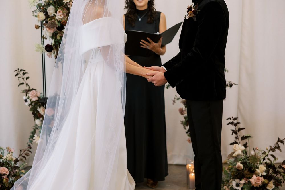 Bride and groom holding hands at altar with officiant during elegant wedding ceremony with floral arrangements.