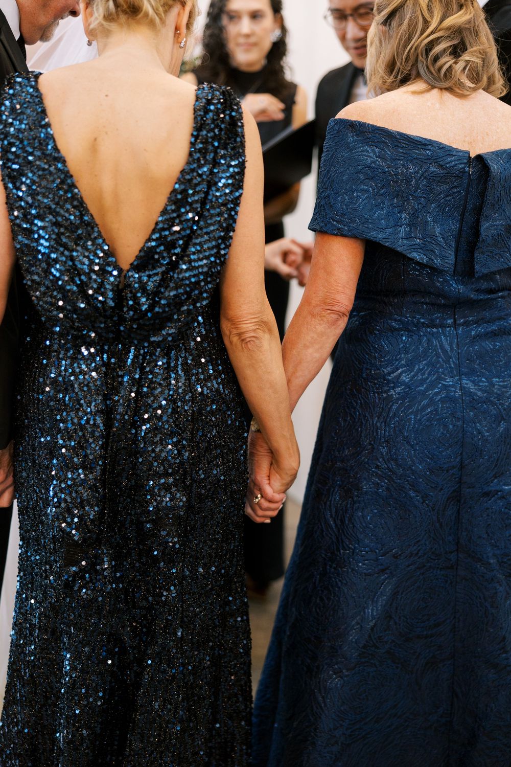 Two women in elegant navy blue gowns hold hands at a formal event, one wearing a stunning backless sequin dress.