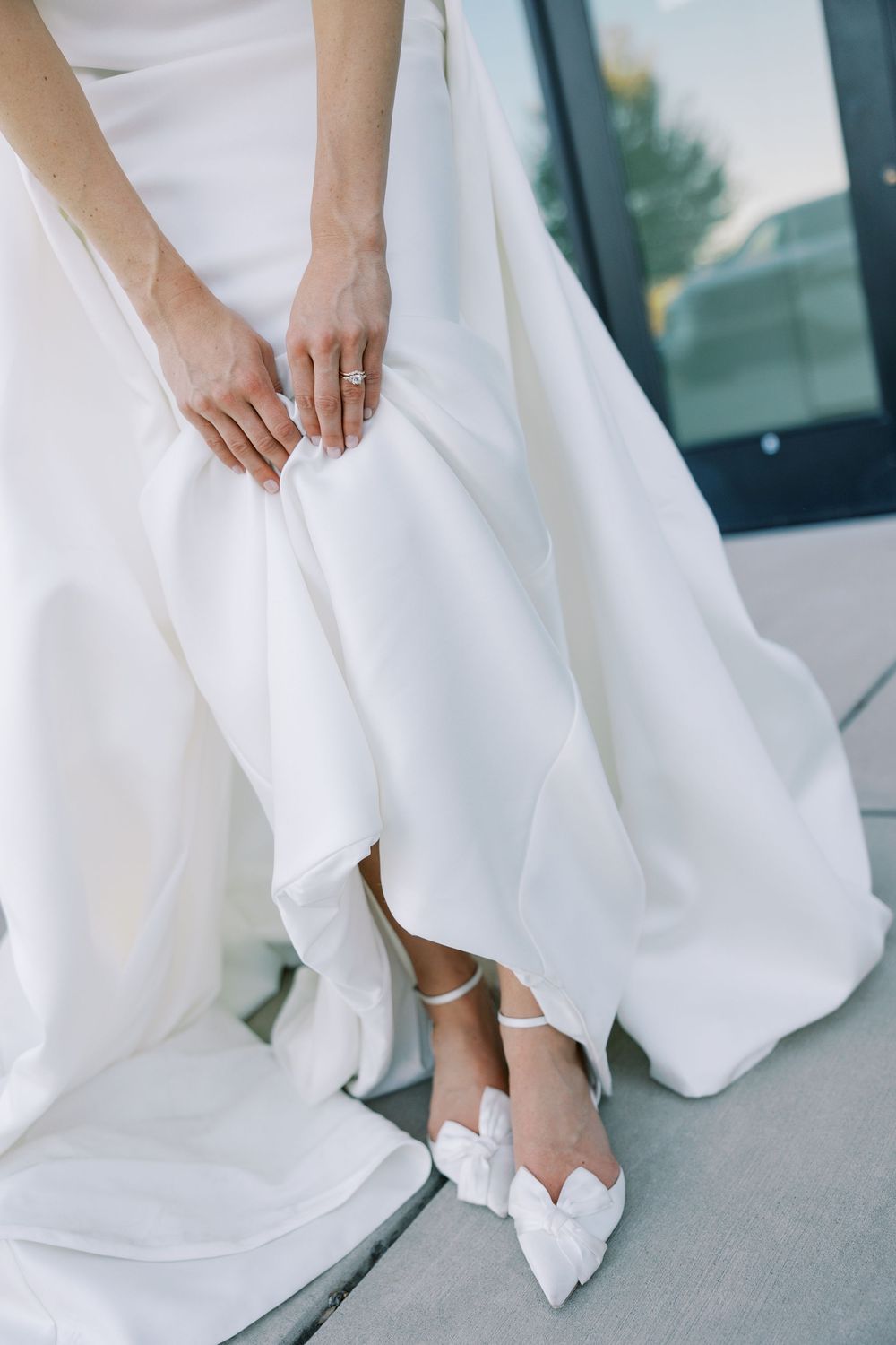 Bride in white satin gown adjusting dress hem wearing white bow-accent heeled wedding shoes.