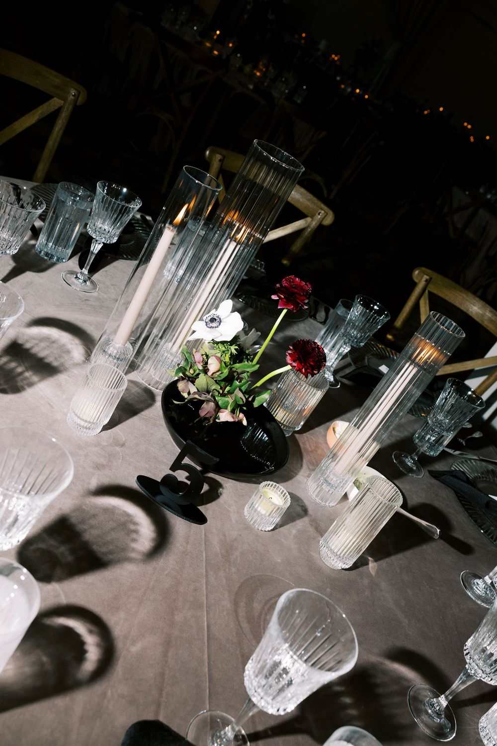Elegant wedding reception table with crystal glassware, tall candles, and dark floral centerpiece on gray linen.