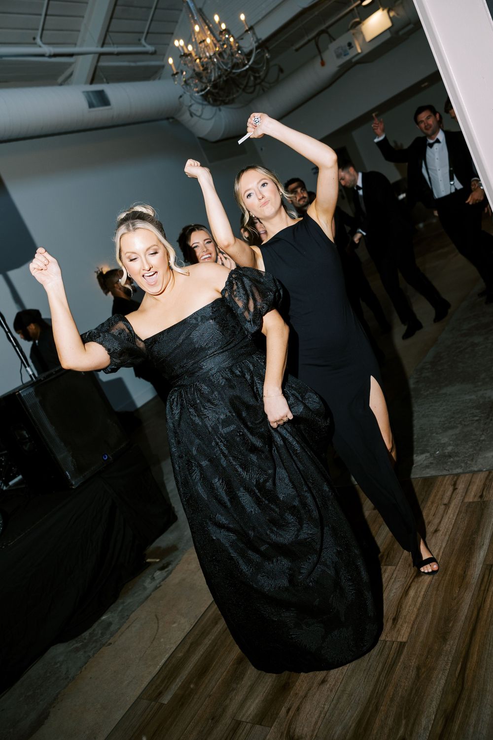 Two women in elegant black gowns dance joyfully at a formal wedding reception on a hardwood dance floor.