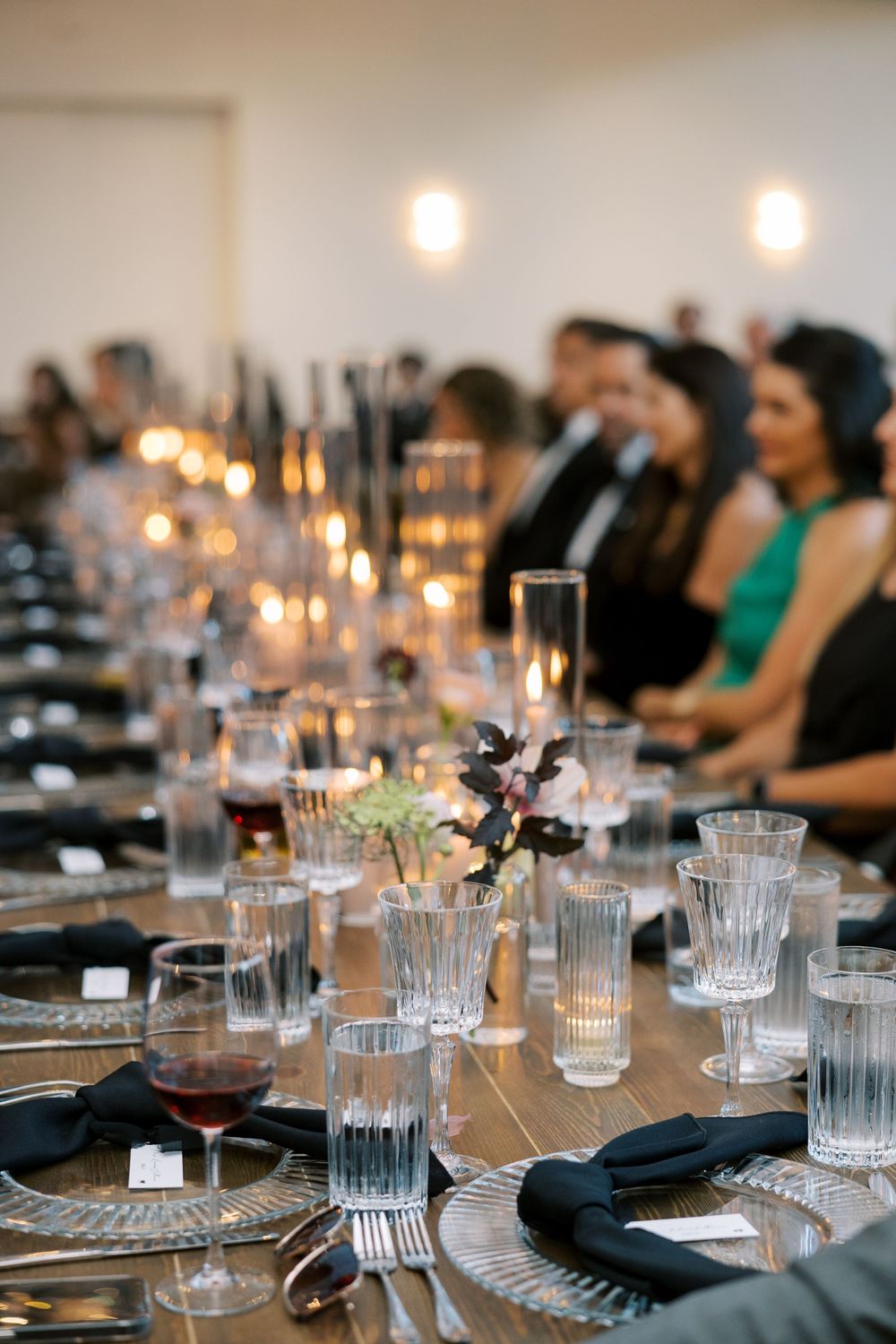 Elegant wedding reception table set with crystal glassware, black napkins, candles, and floral centerpieces with guests in background.
