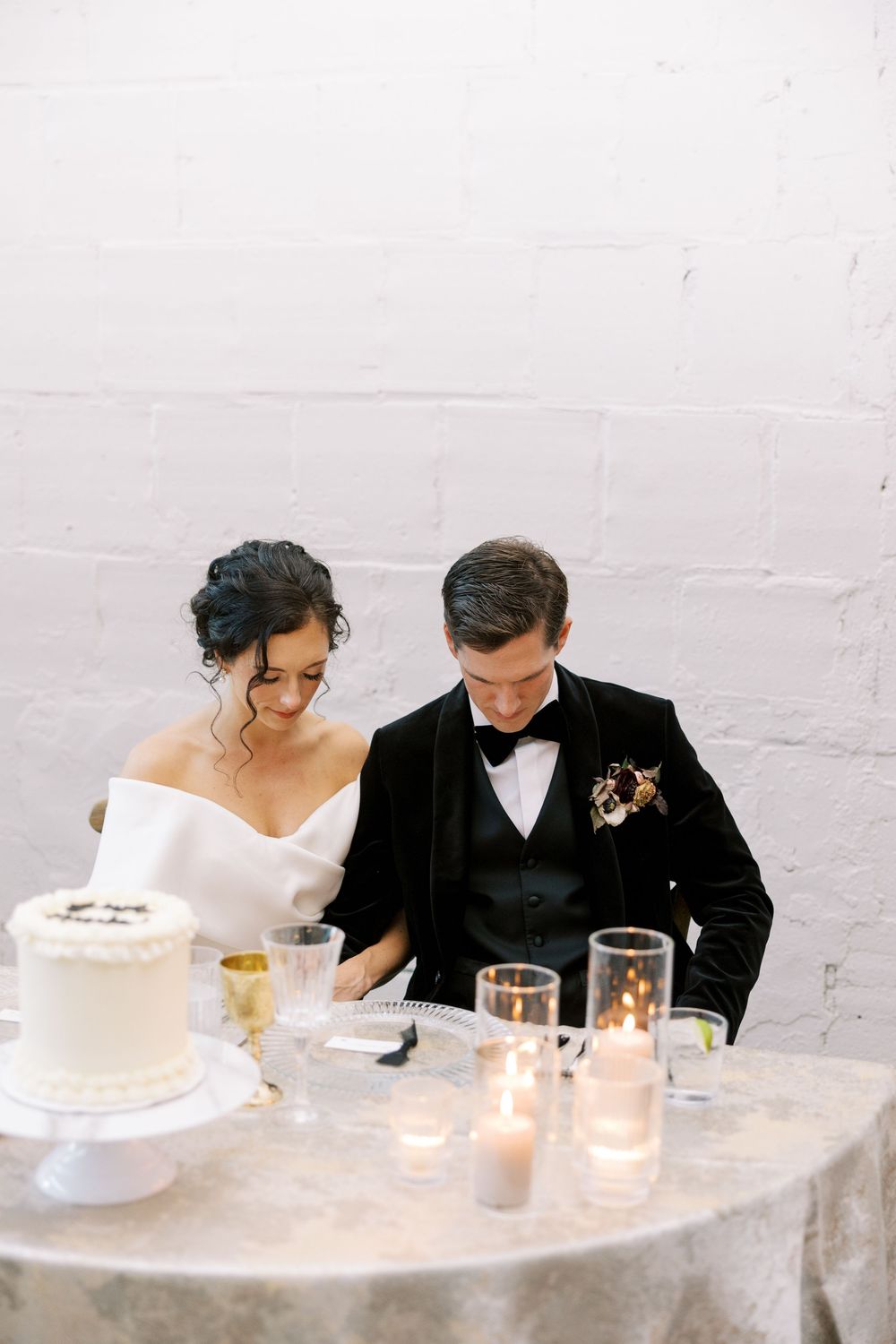 Bride and groom cutting wedding cake at candlelit reception table, white brick wall backdrop.