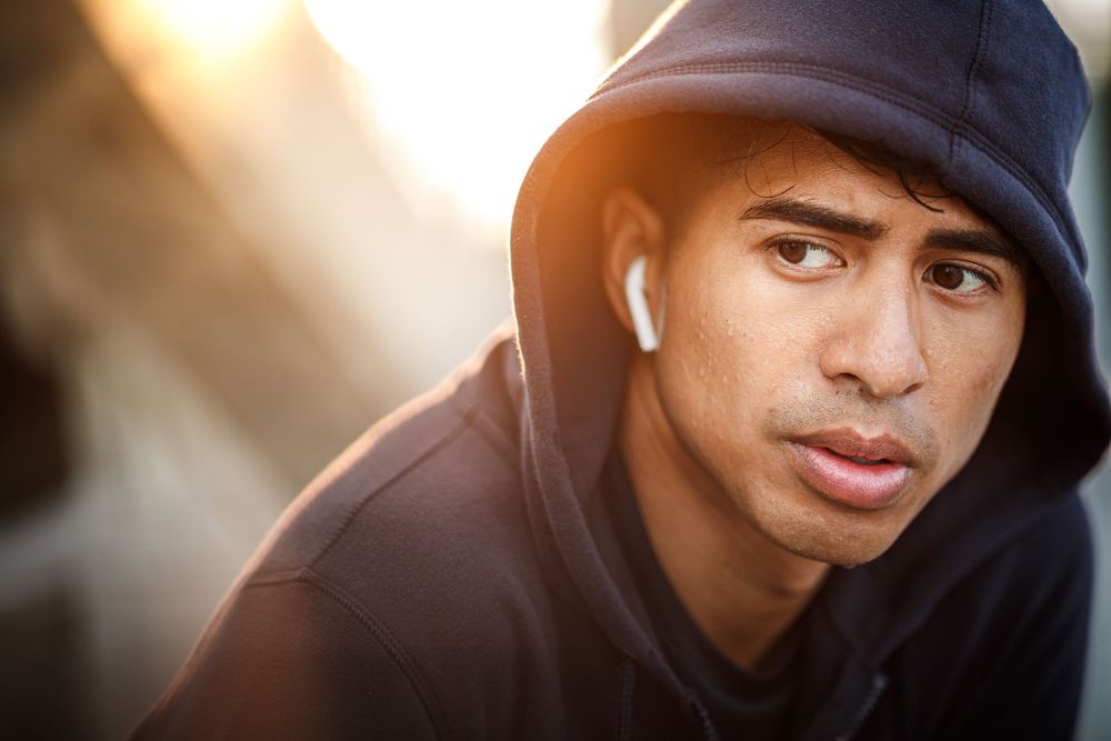 Young man in blue hoodie with AirPods listening to music outdoors in golden sunlight.