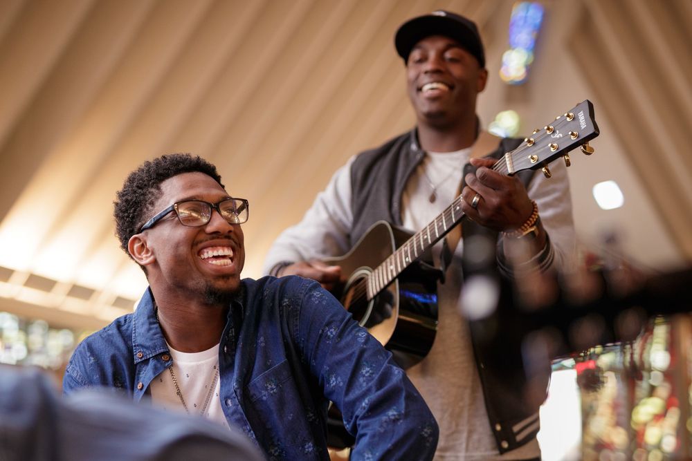 Two men smiling and playing acoustic guitar together indoors, enjoying a fun musical session.