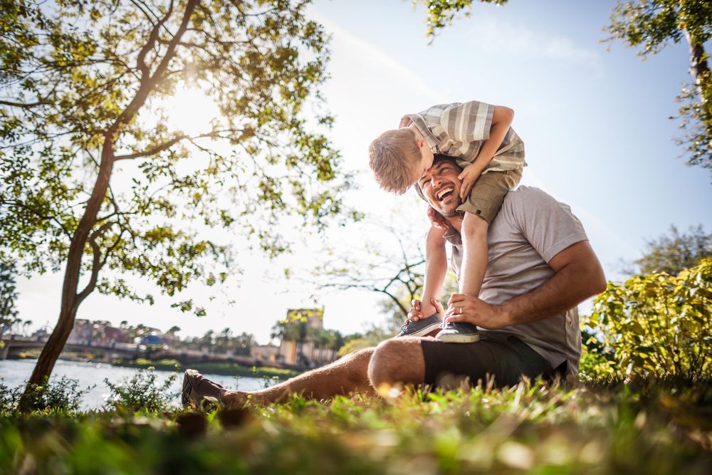 A grandfather and young grandson share a tender moment sitting on grass by a sunlit lake under leafy trees.