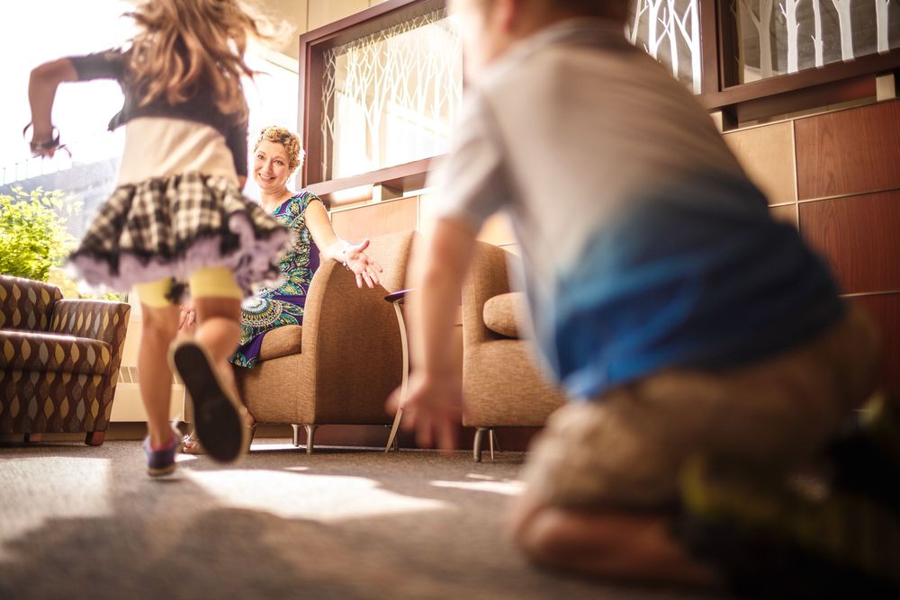 Children running toward a smiling man sitting on a sofa in a bright living room.