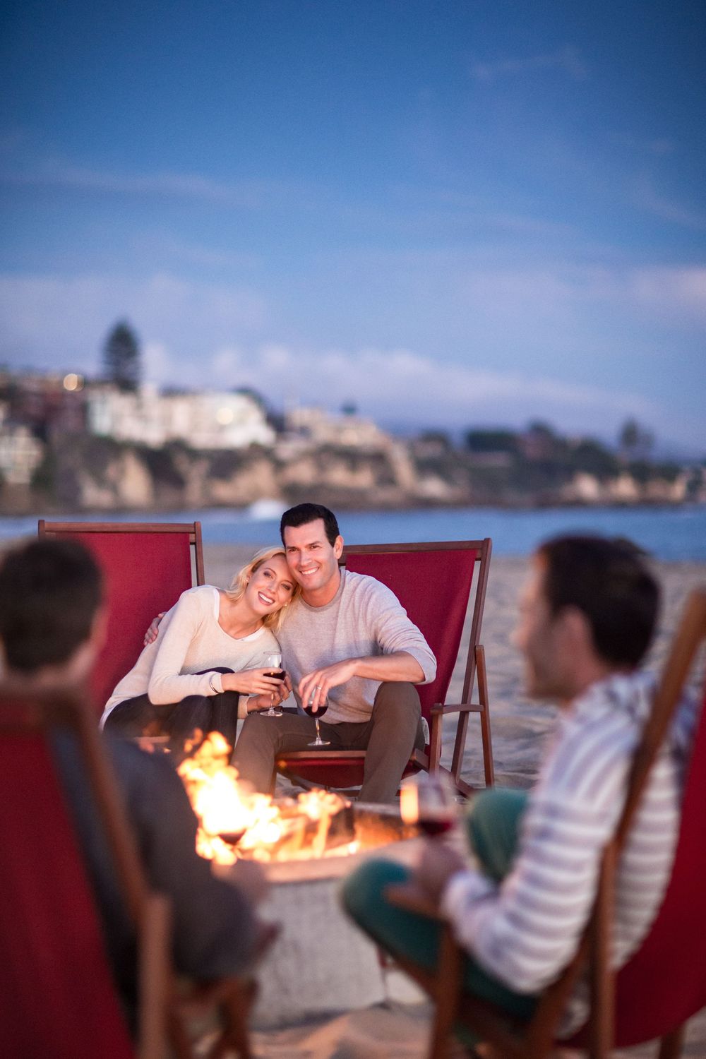 Couples enjoying wine by a beach bonfire at dusk with coastal town in background.