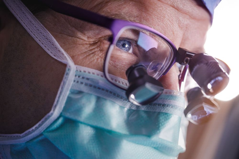 Close-up of a surgeon wearing purple glasses, surgical loupes, and a teal face mask during a medical procedure.