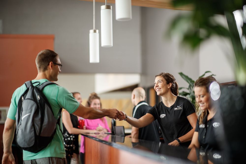A smiling gym receptionist shakes hands with a male member wearing a backpack at a modern fitness center front desk.