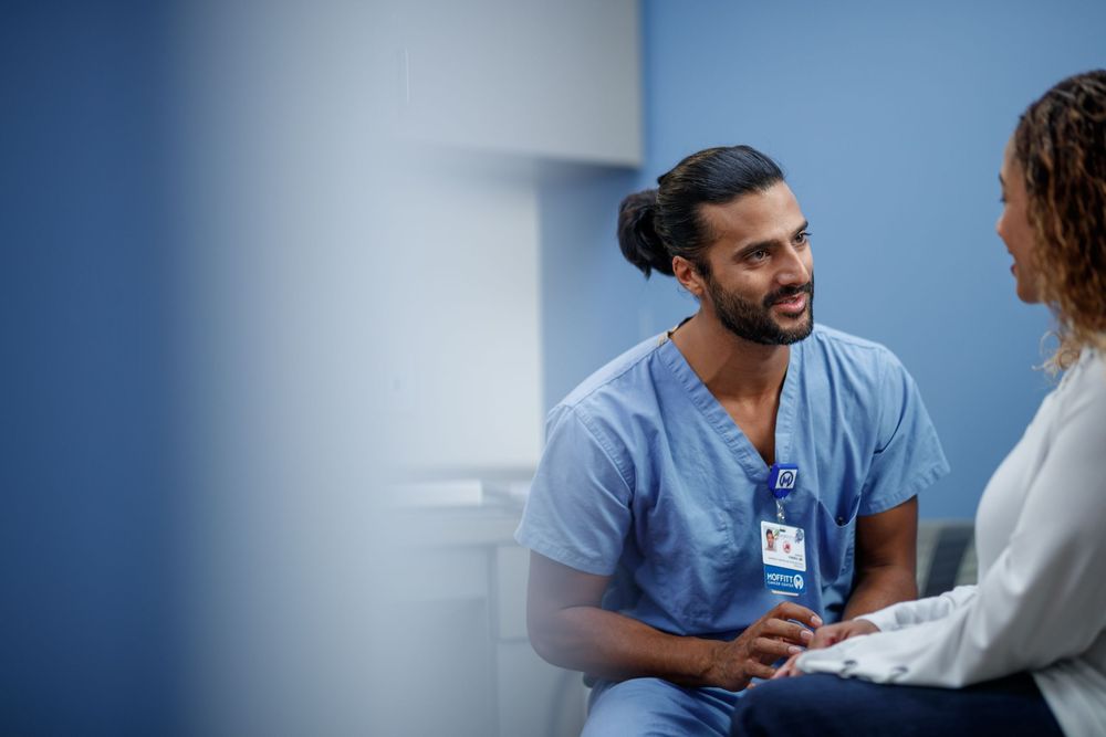 A male nurse in blue scrubs smiles warmly while speaking with a patient in a modern medical facility.