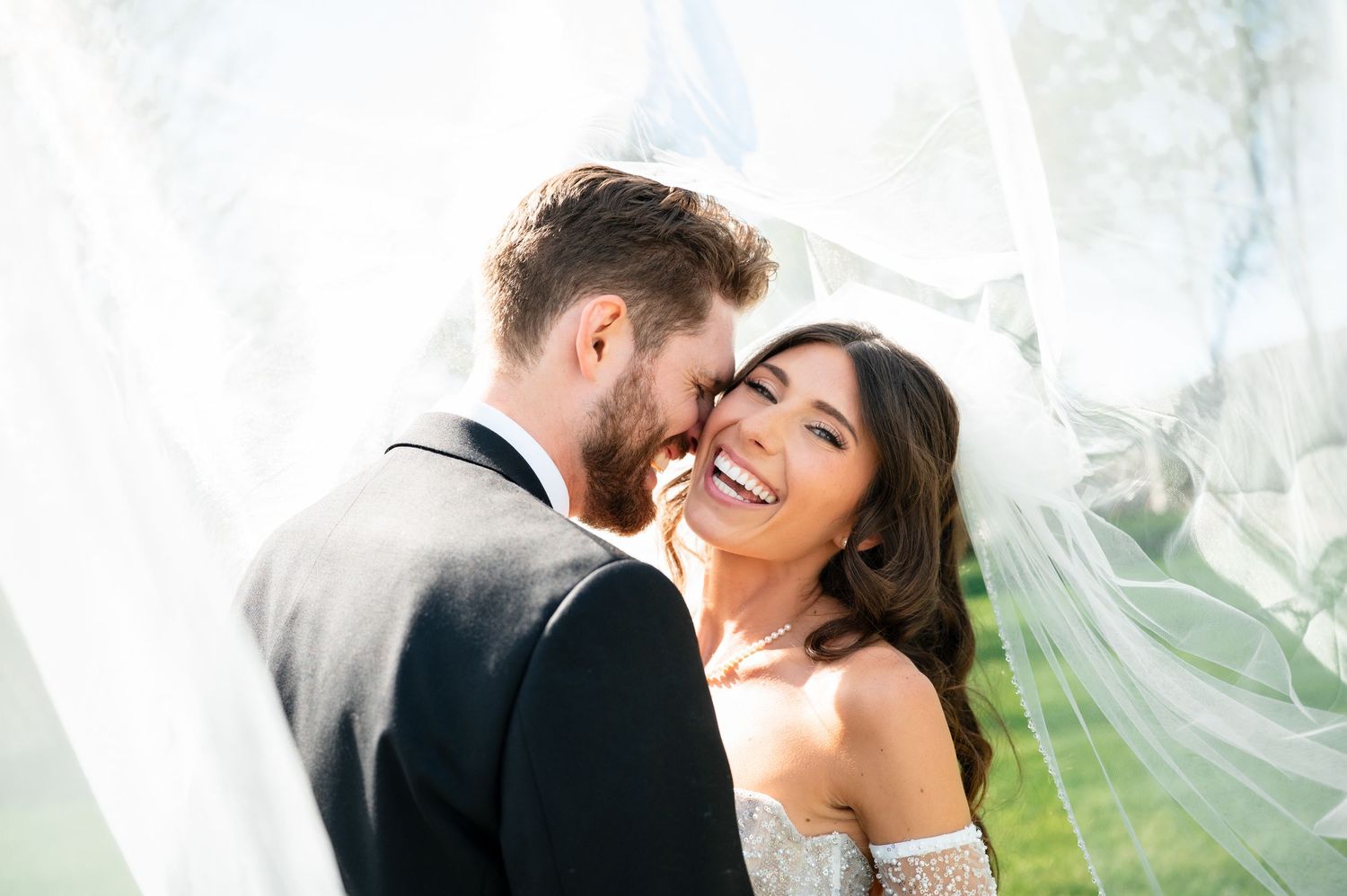 A joyful bride smiles as her groom whispers in her ear beneath a flowing white veil on their wedding day.
