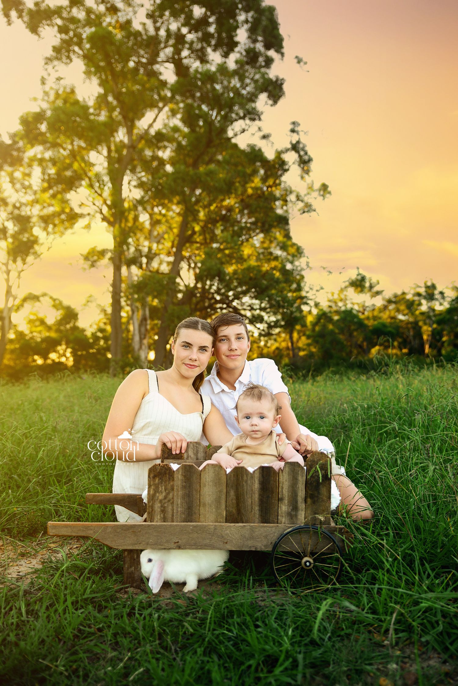 Young family with baby sitting in rustic wooden cart at golden sunset with white rabbit nearby.