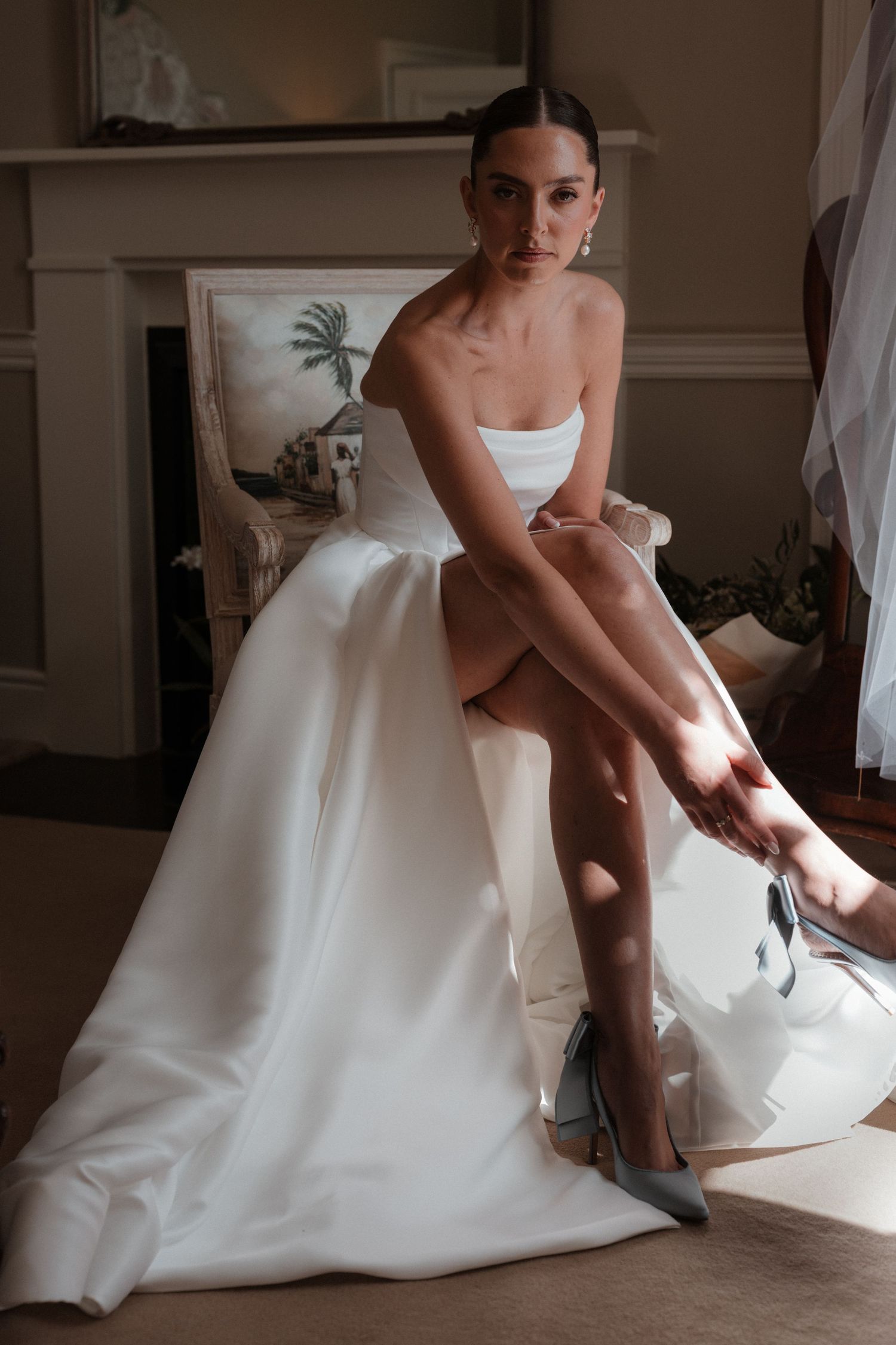 Bride in strapless white ball gown sits elegantly in chair, putting on gray heels, with soft natural light.